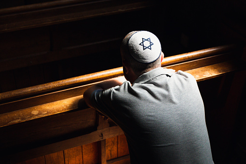 Close up image depicting a young caucasian Jewish adult man in his 30s inside a synagogue. He has his head bowed in prayer and he is wearing the traditional Jewish skull cap - otherwise known as a kippah or yarmulke - on his head. The man has a beard and the background of the synagogue is blurred out of focus. Horizontal color image with copy space.