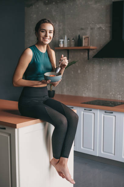 Young lady in sportswear sitting on kitchen table with a bowl of cornflakes and and holding a spoon into her hand while looking at camera. Natural organic nutrition. Healthy lifestyle. stock photo