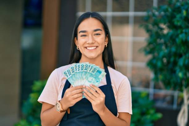 Young latin shopkeeper girl smiling happy holding brazilian real banknotes at the city stock photo