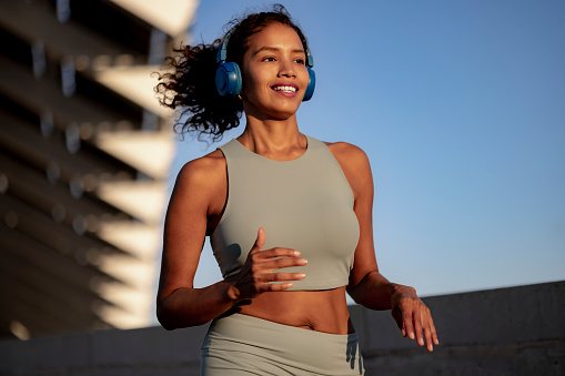 Happy fit Latina woman jogging by herself outdoors, following her daily physical activity routine while listening to music on her wireless headphones