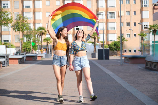 Young lesbian couple smiling, holding hands and walking with a rainbow flag. stock photo