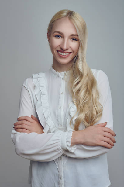 Young long-haired blond woman with her arms folded Young and beautiful long-haired blond woman in white blouse standing with her arms folded and looking at camera with a smirk. Front half-length portrait against grey background application form photos stock pictures, royalty-free photos & images