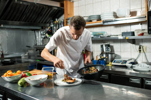 Young male chef with frying pan putting stew on plate with piece of fried salmon stock photo