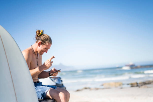 Young male surfer with fingers crossed gambling online using phone at the beach stock photo