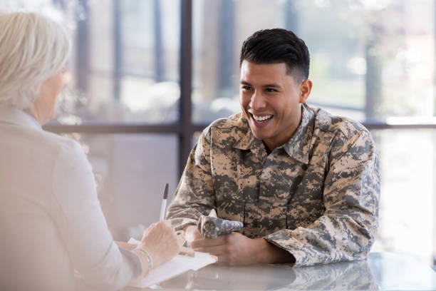 Young male talks with a female counselor and smiles Young, mid-adult male in military uniform sits at a table with mature, female counselor. He smiles at her as she takes notes on their conversation. military lifestyle stock pictures, royalty-free photos & images