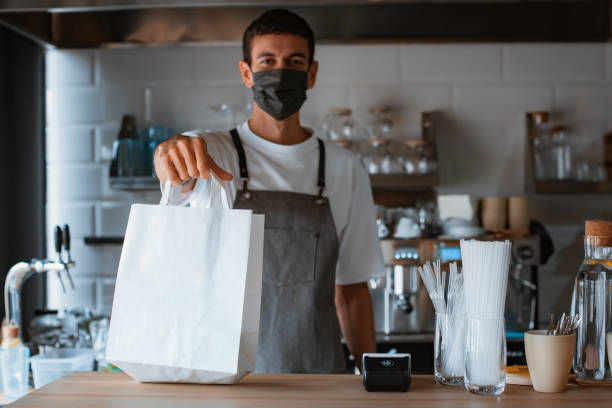 Young man barista in face mask and apron giving pos terminal to customer in coffee shop stock photo