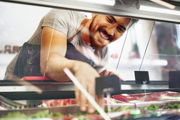 Young man butcher arranging meat products in display case of butcher shop stock photo