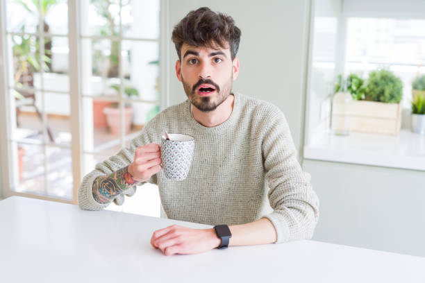 Young man drinking a cup of coffee in the morning scared in shock with a surprise face, afraid and excited with fear expression Young man drinking a cup of coffee in the morning scared in shock with a surprise face, afraid and excited with fear expression worried man funny stock pictures, royalty-free photos & images