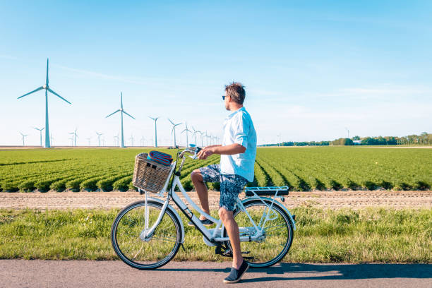 jonge man elektrische groene fiets fiets door windmolenboerderij , windmolens geïsoleerd op een mooie heldere dag nederland flevoland noordoostpolder - zomer nederland stockfoto's en -beelden
