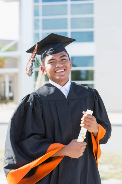 Young man graduating from high school or university A young man, 18 years old, of Asian and  Pacific Islander ethnicity, standing outside a school building wearing a graduation cap and gown. males photos stock pictures, royalty-free photos & images