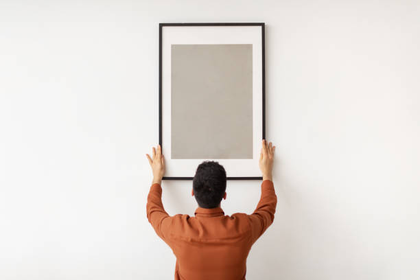 Young man hanging picture frame on the wall Modern Home Interior And Domestic Decor. Rear back view of man hanging painting, putting photo picture frame on white wall. Casual guy holding showing empty mock up poster, blank free copy space one person photos stock pictures, royalty-free photos & images