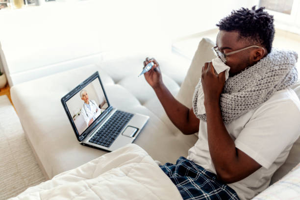 Young man having a video conference with his doctor. Young man getting online medical help and advice during video call with his female doctor and checking symptoms. Ill young man lying down at home under blanket and blowing his nose in one hand showing thermometer to his doctor via laptop. doctor of physiotherapy online stock pictures, royalty-free photos & images