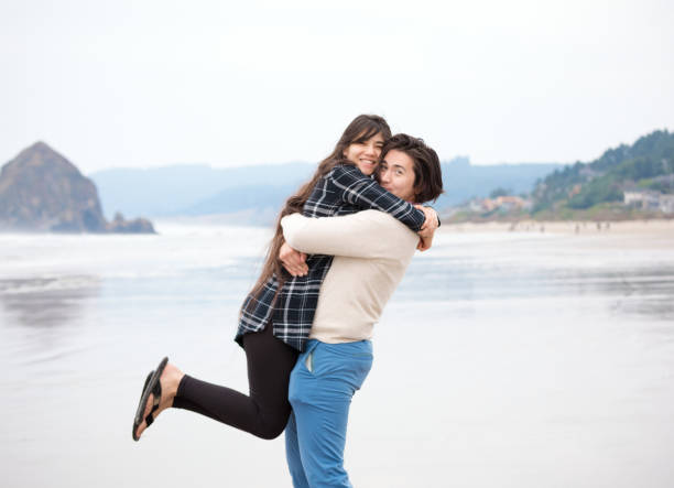 Young man holding smiling woman up on beach by ocean stock photo