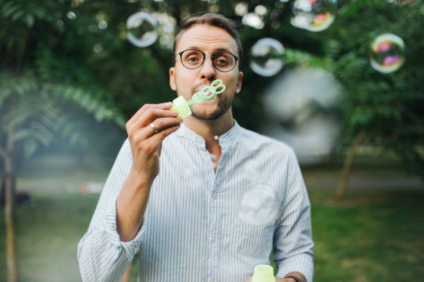 Young man in glasses playing with bubble wands in park stock photo