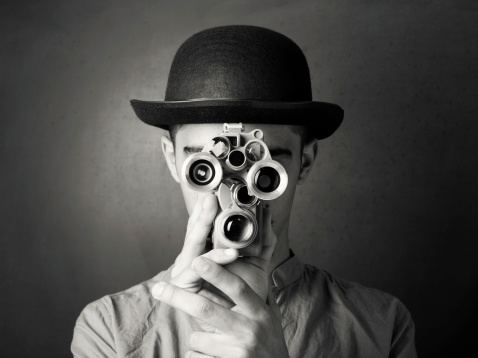Young man in old fashioned style filming via antique camera.The model is wearing bowler hat.He is holding camera in a position that it covers his face.The lenses of camera give eyes look on face.The photo was shot in studio with a full frame DSLR camera in color then edited to black and white.