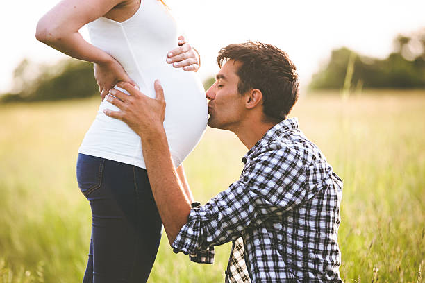young man kissing the belly of his pregnant woman - pregnant couple outside stockfoto's en -beelden