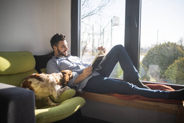 Young man lying on bench next to window and reading book while his dog lying next to him stock photo