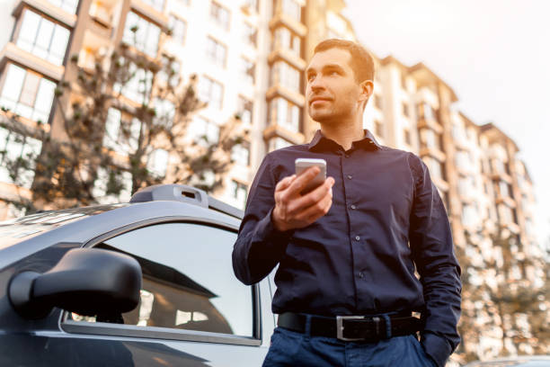 A young man or businessman in a dark shirt stands on the street near the car, looks into the distance in a residential area of the city. The driver is waiting for his passenger or client. A young man or businessman in a dark shirt stands on the street near the car, looks into the distance in a residential area of the city. The driver is waiting for his passenger or client. vlad model photos stock pictures, royalty-free photos & images
