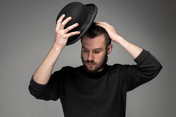 Young man raising his hat  in respect and admiration for Young man with a mustache and beard raising his hat  in respect and admiration for someone. portrait on gray background. male gaze directed downward hats off to you stock pictures, royalty-free photos & images