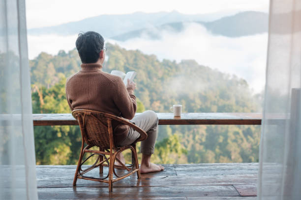 young man reading book near window and looking mountain view at countryside homestay in the morning sunrise. SoloTravel, journey, trip and relaxing concept stock photo