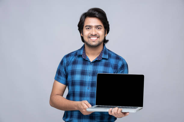young man showing blank screen of laptop on gray background:- stock photo stock photo