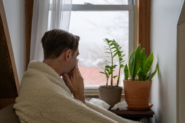 A young man sick at home uses tissues by a window on a rainy day. stock photo