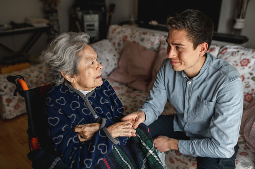 Young man sitting next to an old sick aged woman in wheelchair taking her hands while talking and smiling. Family, home care concept.