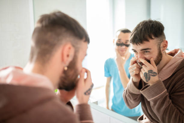Young man squeezing blackheads on nose, his boyfriend wearing facial mask Young man standing in front of the bathroom mirror and squeezing blackheads on his nose, his boyfriend wearing facial mask blackheads stock pictures, royalty-free photos & images