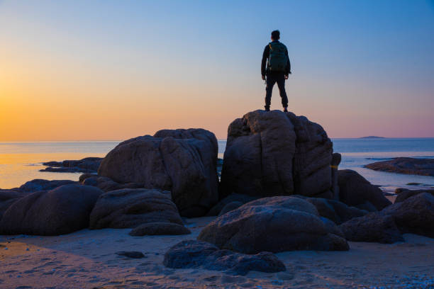 Young man standing on rock looking at sea sunrise stock photo