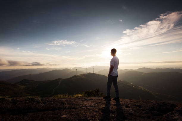 Young man standing on top of cliff in summer mountains at sunrise stock photo