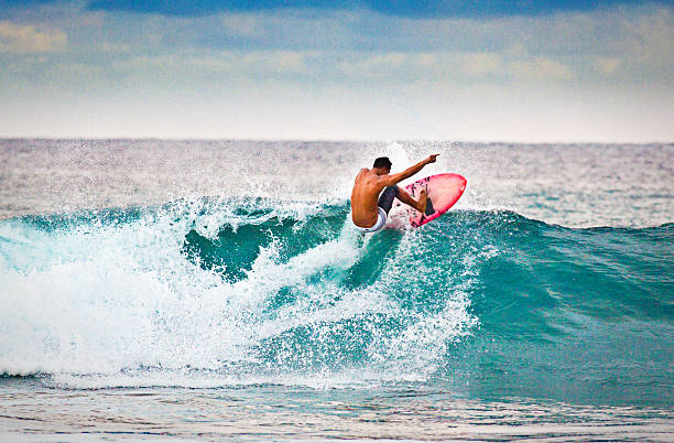 Young Man surfer Surfing in the Beach of Kauai, Hawaii A young native Hawaii man surfing on the wave of Poipu Beach, on the island of Kauai, Hawaii, USA. He is on a pink surfboard and making a turn on the wave in the aqua sea of Kauai. Photographed in horizontal format with copy space available. big wave surfing stock pictures, royalty-free photos & images