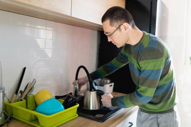 Young man turning on the faucet to fill the kettle up with water by the kitchen sink stock photo