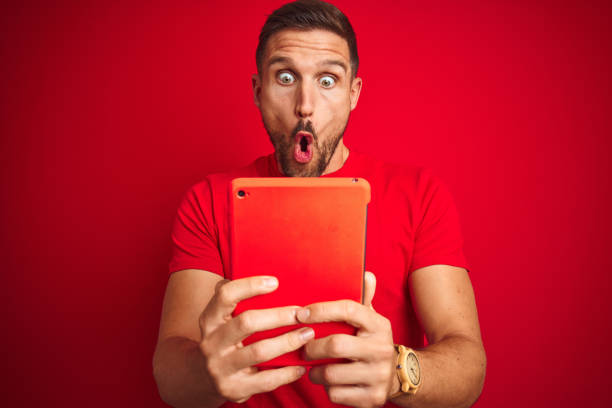 Young man using digital touchpad tablet over red isolated background scared in shock with a surprise face, afraid and excited with fear expression Young man using digital touchpad tablet over red isolated background scared in shock with a surprise face, afraid and excited with fear expression worried man funny stock pictures, royalty-free photos & images