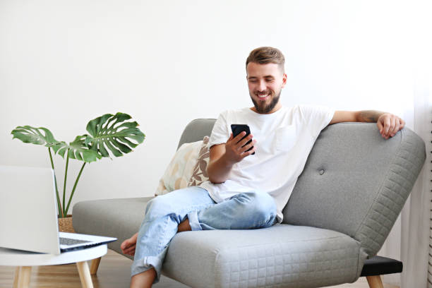Young man with full beard chilling in his apatrment. Bearded guy wearing blank white t-shirt & denim pants sitting alone at home on grey textile couch. Young man w/ facial hair in domestic situations. Interior background, copy space, close up, monstera. portability stock pictures, royalty-free photos & images