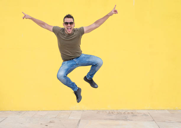 Young man with sunglasses jumping in front of a yellow wall. stock photo
