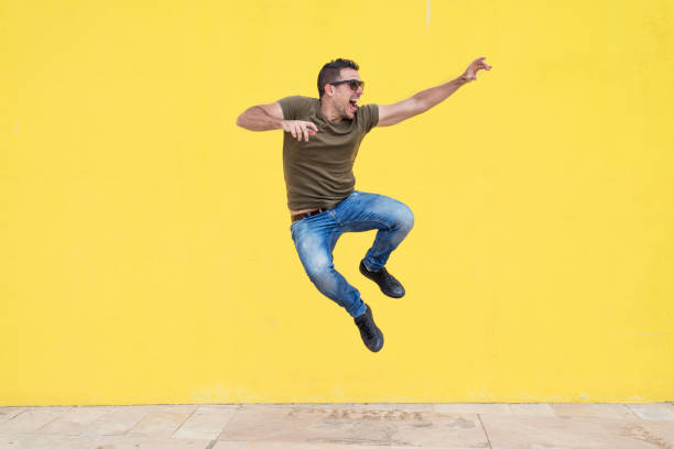 Young man with sunglasses jumping in front of a yellow wall. stock photo
