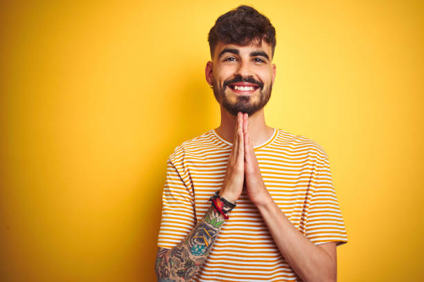 Young man with tattoo wearing striped t-shirt standing over isolated yellow background praying with hands together asking for forgiveness smiling confident.  prayer request stock pictures, royalty-free photos & images
