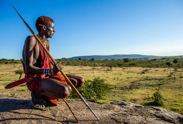 Young Masai warrior is sitting on a big stone in traditional clothing with a spear in the savannah against a blue sky. Tanzania, East Africa - august 12, 2018: Young Masai warrior is sitting on a big stone in traditional clothing with a spear in the savannah against a blue sky. Tanzania, East Africa, August 12, 2018. maasai warrior stock pictures, royalty-free photos & images
