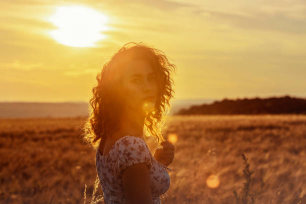 young moroccan woman, with brown curly hair, standing in a wheat field, while the sun ist setting in the background and blinding the camera stock photo