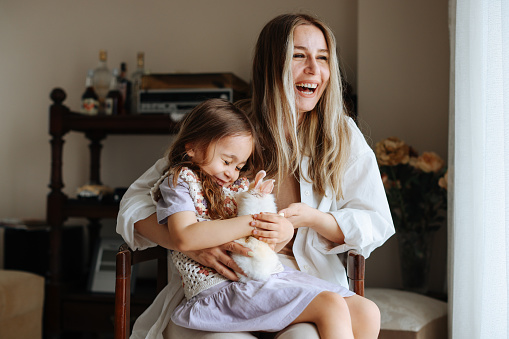 Shot of a mother and daughter hugging their pet rabbit at home. Young mother holding furry cute baby pet rabbit showing it to her toddler daughter indoors.