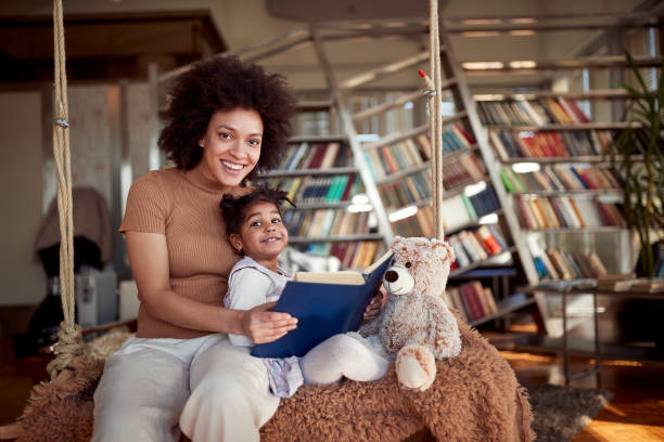 A young mother and her little daughter are posing while reading a book sitting on the swing at home together. Family, together, leisure stock photo