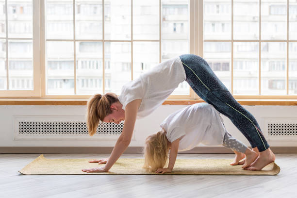 young mother doing yoga with 3-years girl in front of window. downward facing dog asana - yoga crianças imagens e fotografias de stock