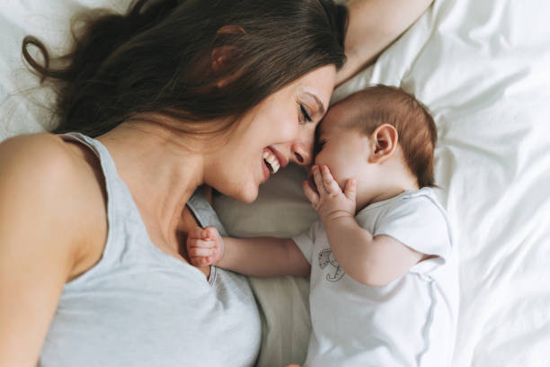 jonge moeder die pret met leuk babymeisje op het bed, natuurlijke tonen, liefdeemotie heeft - moeder stockfoto's en -beelden