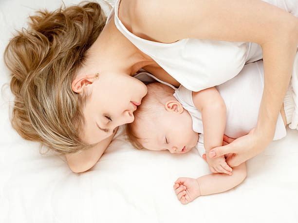 Young mother laying with her baby on white linens stock photo