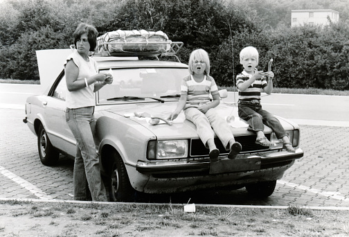 Vintage monochrome 1978 image; young mother with daugter and son sitting on hood of car eathing lunch sausages on a roadtrip holiday in Germany.