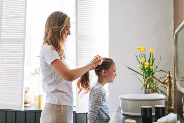 Young mother woman with long hair with little tween girl daughter in pajamas combs her hair on morning in bathroom at home stock photo