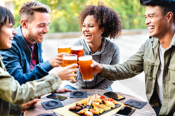 Young multicultural friends drinking and toasting beer at brewery bar patio - Youth life style concept with men and women having fun together out side - Bright vivid filter with focus on mid girl stock photo