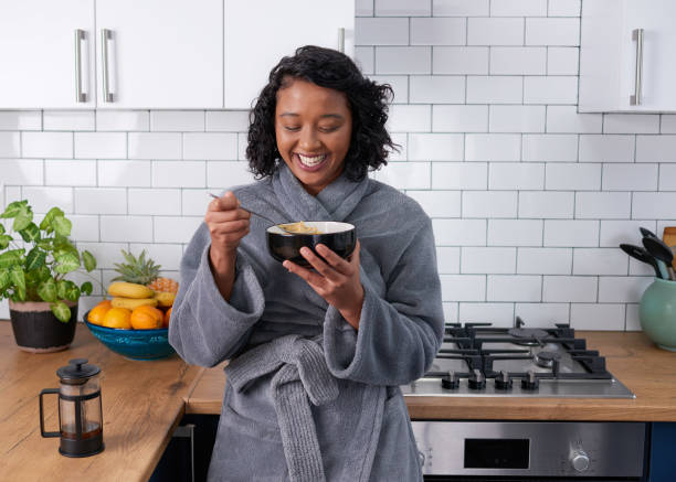 A young multi-ethnic woman laughs while eating breakfast cereal in dressing gown stock photo