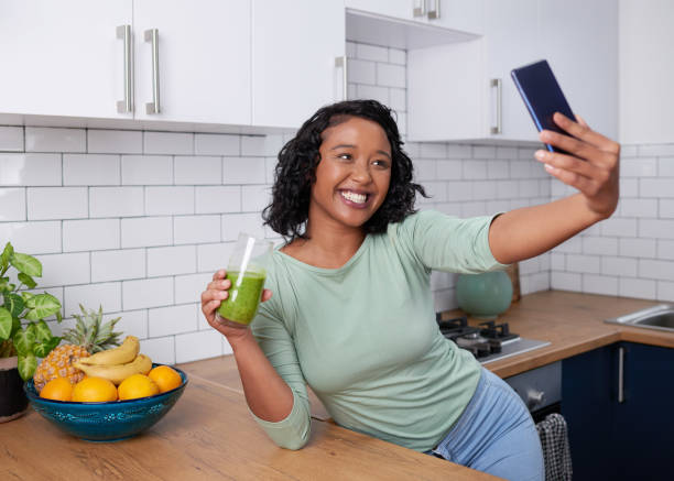 A young multiracial woman takes a selfie with her green smoothie in the kitchen stock photo