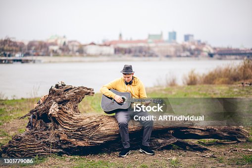 istock Young musician teen boy sitting on a tree trunk near the river with Warsaw panorama behind, playing, practicing his guitar, being focused, professional, making effort. 1301779086